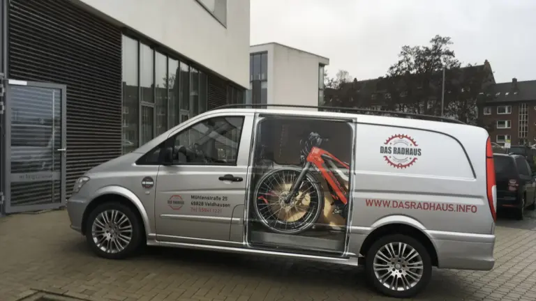 Silberner Transporter mit offener Schiebetür, Fahrrad im Laderaum, Logo und Webadresse auf der Seite