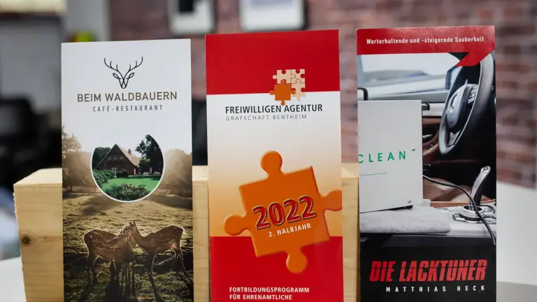 Vier gedruckte Flyer nebeneinander auf einem Tisch vor einer Ziegelwand